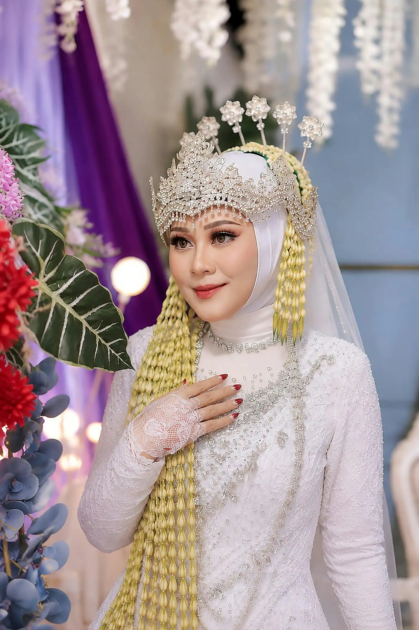 A bride in a white hijab with a gold jewelled headpiece and long jasmine strands, hand on her chest, defined eyes and rosy lips