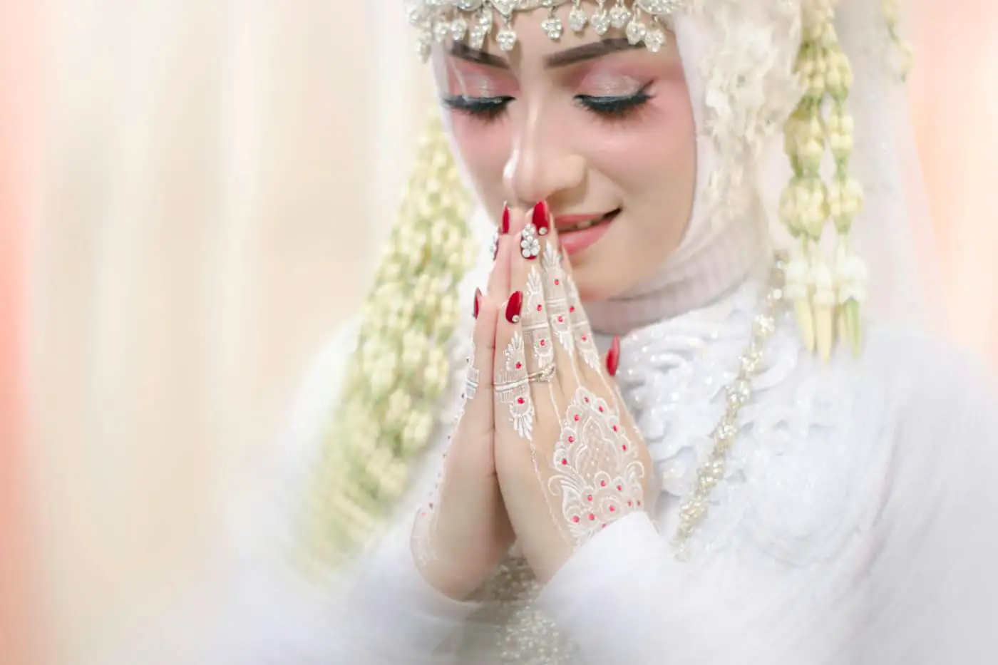 A smiling bride with closed eyes pressing her henna-decorated hands together, in a white hijab with jasmine strands and a jewelled headpiece