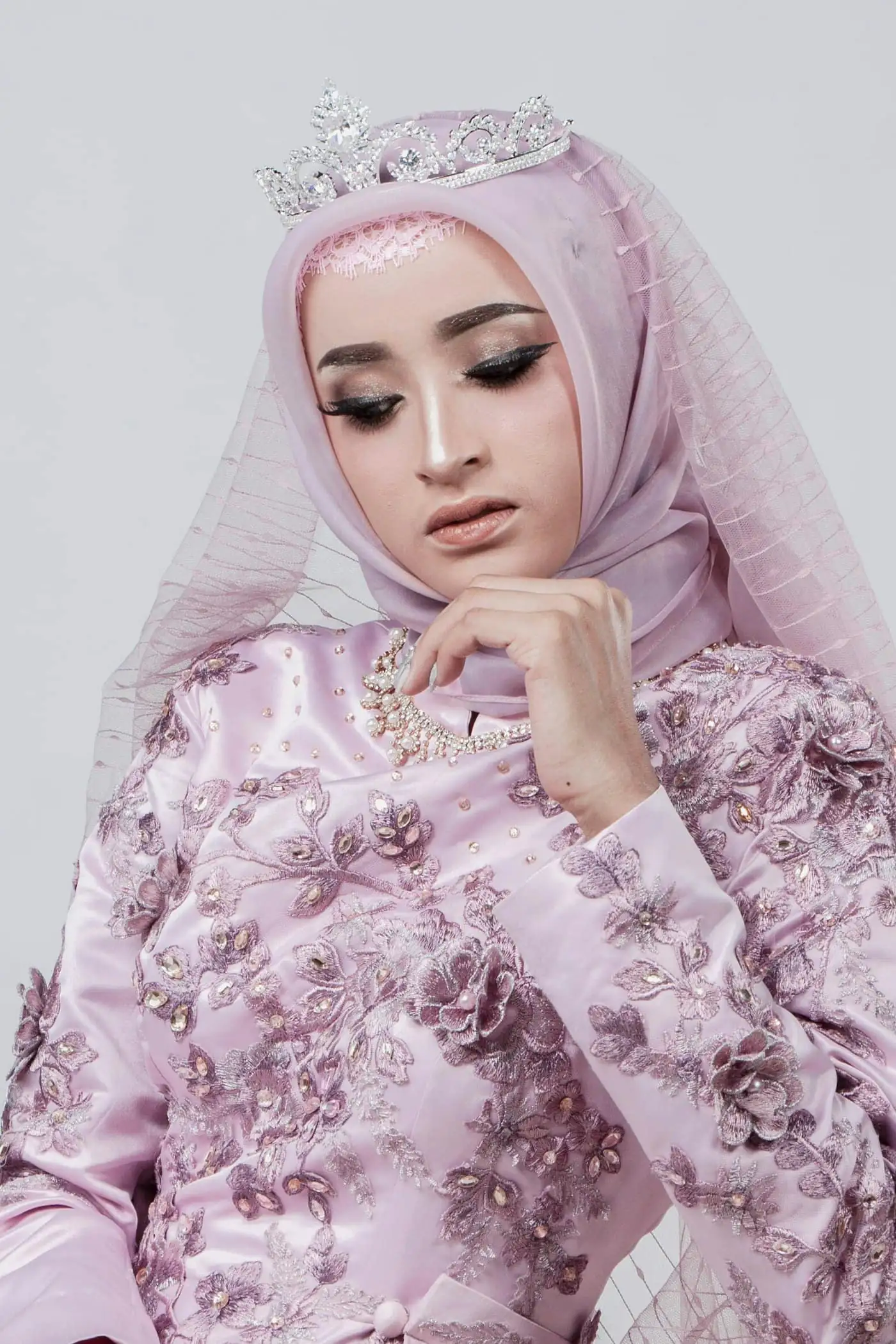 A woman in a pink hijab with a light veil and silver tiara, and a pink lace kebaya, resting her hand under her chin and looking down