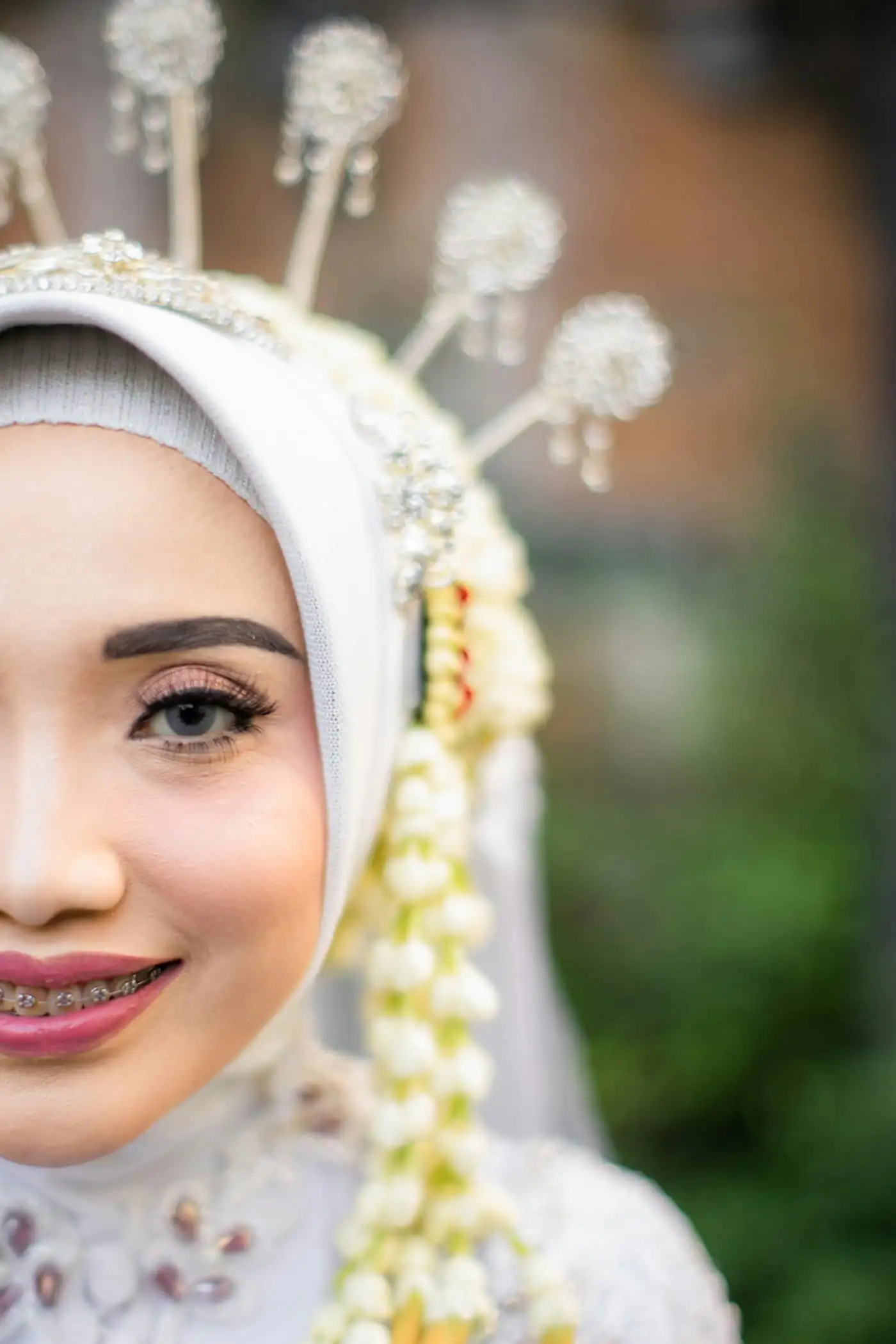 Close-up of the bride’s smiling face, half in frame, in a white hijab with a gold crown-style headpiece, jewelled pins and jasmine strands