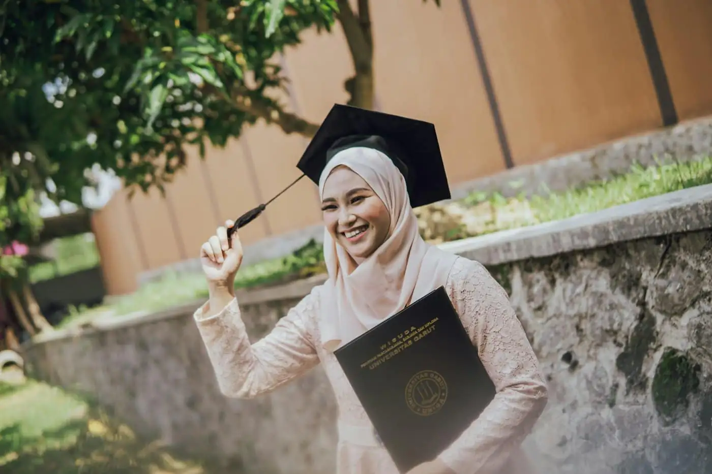 A smiling graduate in a cream hijab and mortarboard holding her black certificate folder, outdoors beside a stone wall