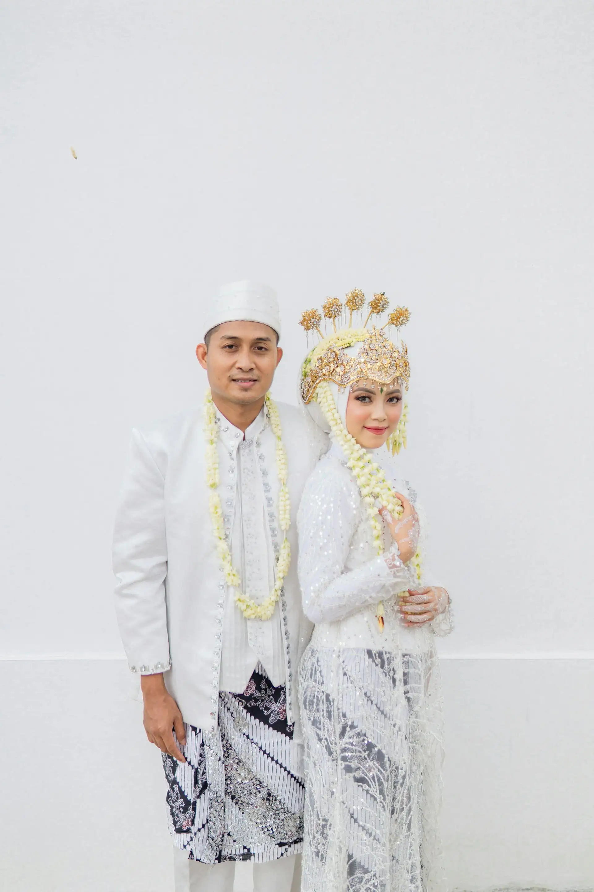 A groom in a white baju and songkok and a bride in white lace with a gold crown-style headpiece, both wearing jasmine strands, standing side by side against a plain white wall