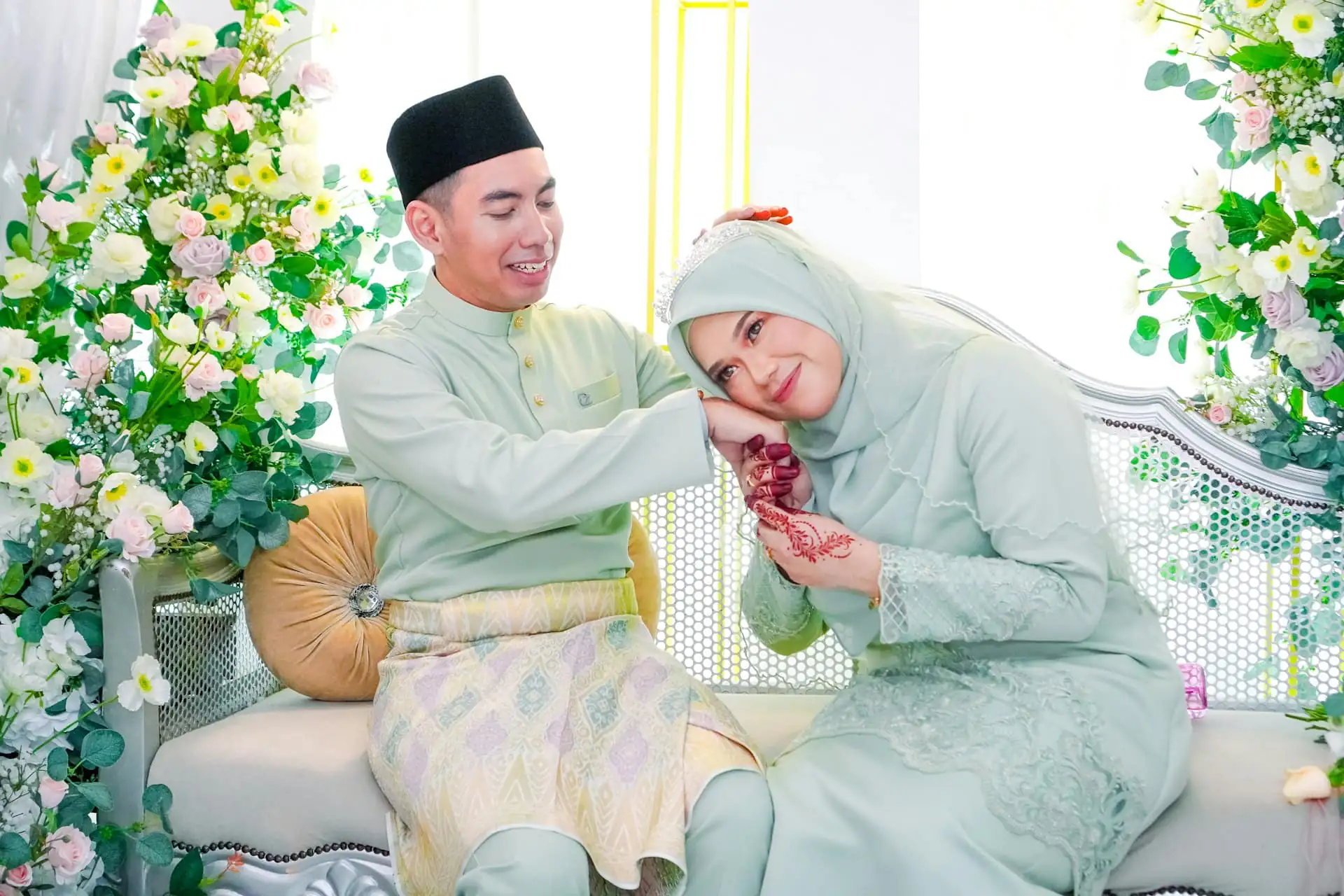 A groom in a mint baju Melayu and songkok and a bride in a mint hijab and lace kurung seated together on a sofa surrounded by pink and white flowers, she resting her cheek on his henna-decorated hand