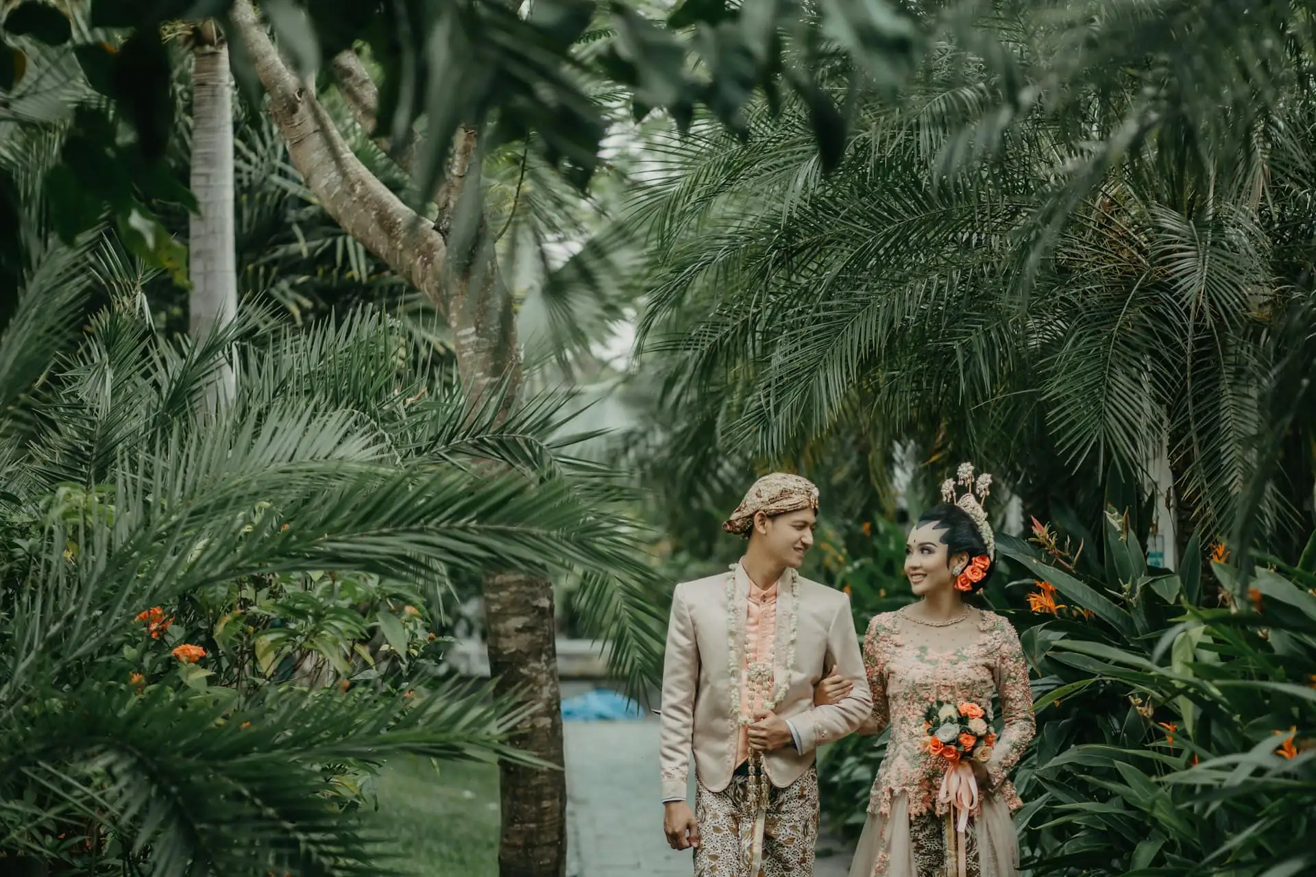 A couple in cream and orange traditional wedding attire walking arm in arm through a lush tropical garden of palms