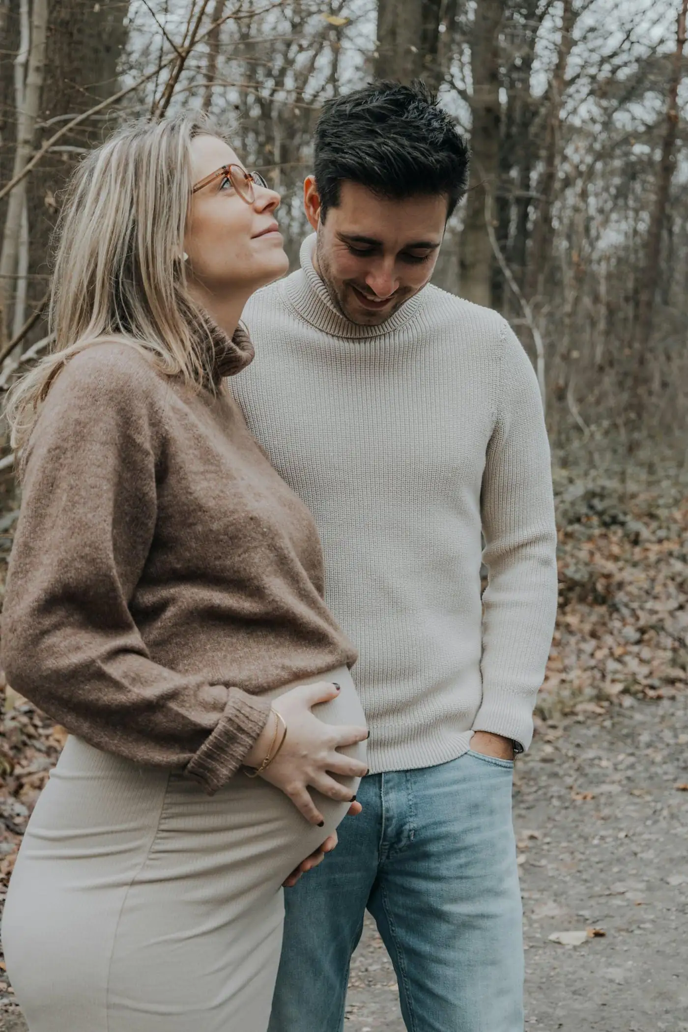 An expecting couple in cream knitwear standing close among bare trees, he looking down at her bump as she looks up