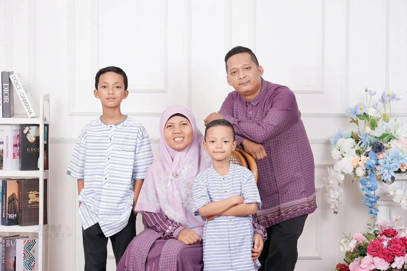 A father, mother and two sons in coordinated lilac and blue-striped Raya outfits posed together in front of a white panelled backdrop with flowers