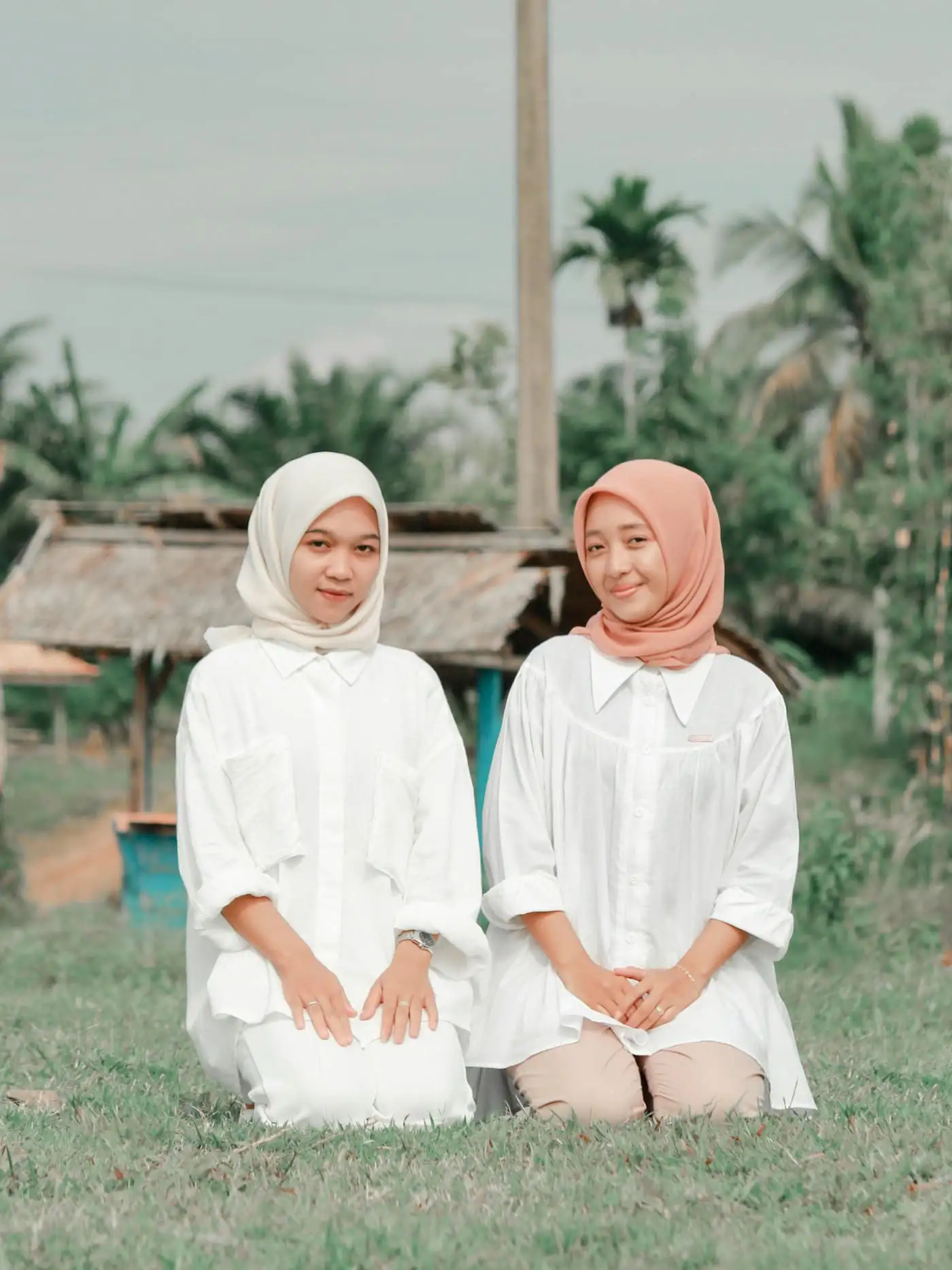 Two young women in white shirts, one in a white hijab and one in a coral hijab, kneeling side by side on a lawn with palm trees behind them